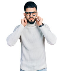 Fototapeta premium Young arab man with beard wearing elegant turtleneck sweater and glasses covering ears with fingers with annoyed expression for the noise of loud music. deaf concept.