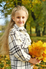 Little blonde girl posing in a white plaid shirt in an autumn park with a bouquet of yellow leaves