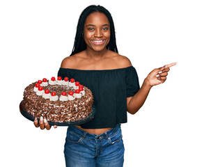 Young african american woman celebrating birthday holding big chocolate cake smiling happy pointing with hand and finger to the side