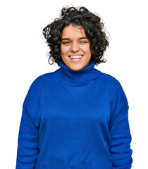 Young hispanic woman with curly hair wearing turtleneck sweater winking looking at the camera with...