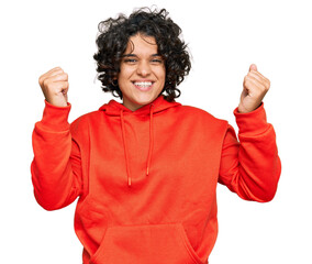 Young hispanic woman with curly hair wearing casual sweatshirt screaming proud, celebrating victory and success very excited with raised arms