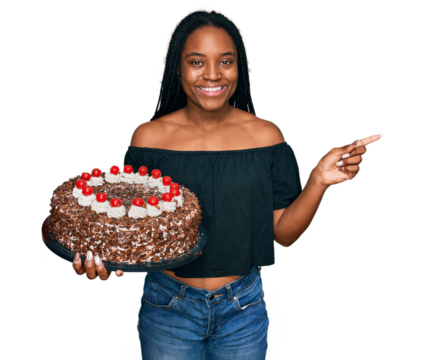 Young african american woman celebrating birthday holding big chocolate cake smiling happy pointing with hand and finger to the side - Powered by Adobe