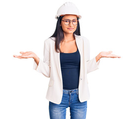 Young beautiful latin girl wearing architect hardhat and glasses clueless and confused expression with arms and hands raised. doubt concept.