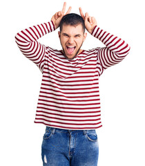 Young handsome man wearing striped sweater posing funny and crazy with fingers on head as bunny ears, smiling cheerful