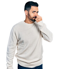 Young hispanic man with beard wearing casual white sweater smelling something stinky and disgusting, intolerable smell, holding breath with fingers on nose. bad smell