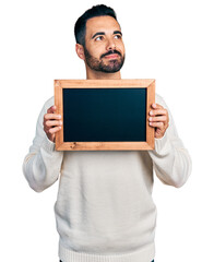 Young hispanic man with beard holding blackboard smiling looking to the side and staring away...
