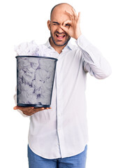 Young handsome man holding paper bin full of crumpled papers smiling happy doing ok sign with hand on eye looking through fingers