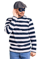 Young handsome man wearing burglar mask smiling with hand over ear listening an hearing to rumor or gossip. deafness concept.