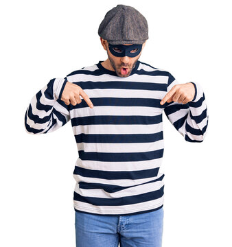Young handsome man wearing burglar mask pointing down with fingers showing advertisement, surprised face and open mouth