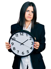 Young brunette woman with blue eyes holding big clock skeptic and nervous, frowning upset because of problem. negative person.