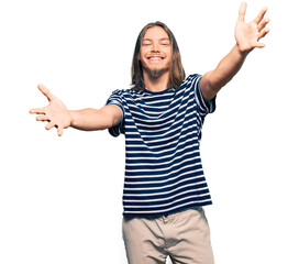 Handsome caucasian man with long hair wearing casual striped t-shirt looking at the camera smiling with open arms for hug. cheerful expression embracing happiness.