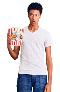 Young african american man holding popcorn scared and amazed with open mouth for surprise, disbelief face