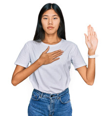 Beautiful young asian woman wearing casual white t shirt swearing with hand on chest and open palm,...