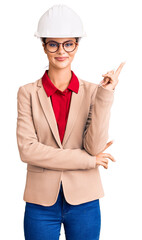 Young beautiful woman wearing architect hardhat smiling happy pointing with hand and finger to the side