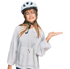 Teenager caucasian girl wearing bike helmet smiling cheerful presenting and pointing with palm of hand looking at the camera.