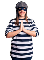 Young beautiful brunette woman wearing burglar mask praying with hands together asking for forgiveness smiling confident.