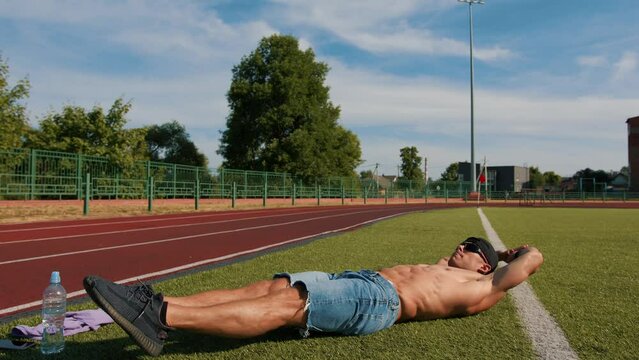 Young muscular man with naked torso is training in city stadium and doing exercise abs fold