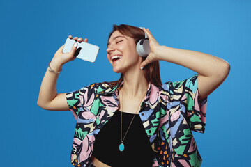 Young student girl with wireless headphones, singing loud in smartphone as microphone, having fun...