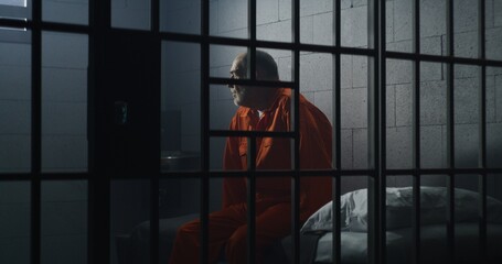 Elderly criminal in orange uniform sits on jail bed and talks with cellmate. Depressed prisoners...