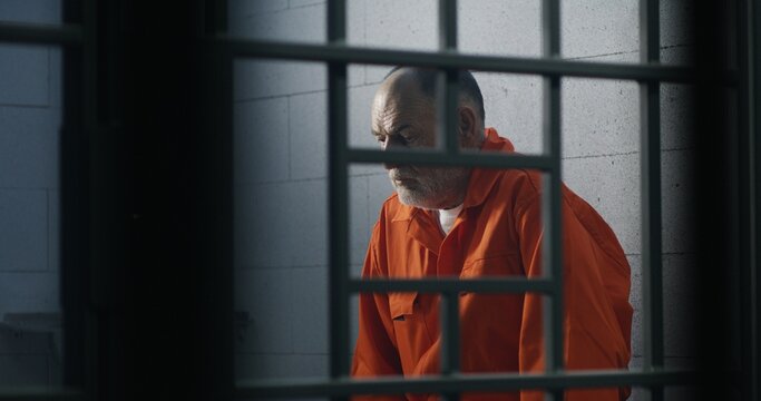 Elderly criminal in orange uniform sits on prison bed and thinks about freedom. Prisoner serves imprisonment term in jail cell. Guilty inmate in detention center or correctional facility.
