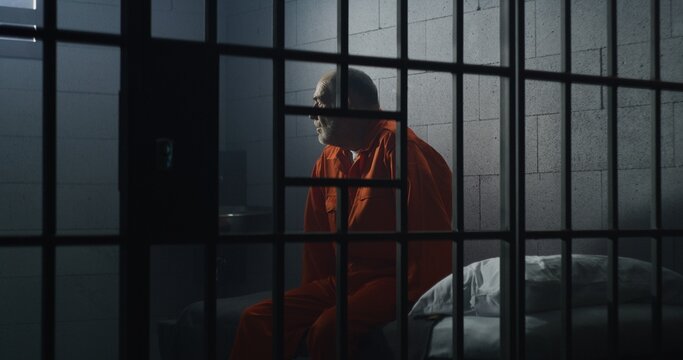 Elderly criminal in orange uniform sits on jail bed and talks with cellmate. Depressed prisoners serve imprisonment term in prison cell. Guilty inmates in detention center or correctional facility.