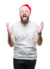 Young caucasian hipster man wearing christmas hat over isolated background crazy and mad shouting and yelling with aggressive expression and arms raised. Frustration concept.