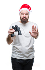 Young caucasian hipster man wearing christmas hat looking though binoculars over isolated background with open hand doing stop sign with serious and confident expression, defense gesture