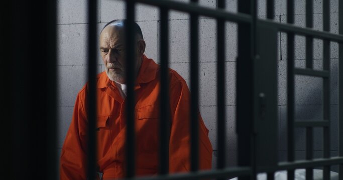 Elderly criminal in orange uniform sits on prison bed and thinks about freedom. Prisoner serves imprisonment term in jail cell. Guilty inmate in detention center or correctional facility.