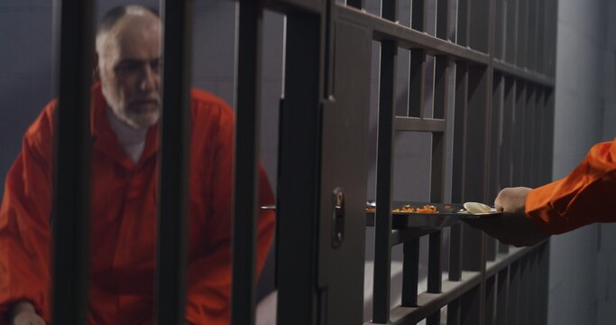 Elderly prisoner in orange uniform sits in prison cell. Prison guard gives him tray of food through metal bars. Guilty criminal serves imprisonment term for crime. Inmate in jail or detention center.