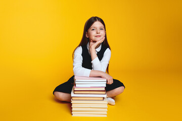 Clever student girl wearing school uniform, holding hand under chin while sitting near pile of books, against yellow background Back to school concept