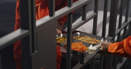 Elderly prisoner in orange uniform sits in prison cell. Prison guard gives him tray of food through...