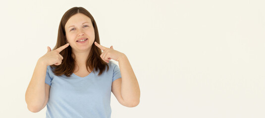 Naklejka na ściany i meble Excited woman wearing t-shirt pointing fingers at her cheek isolated over white background