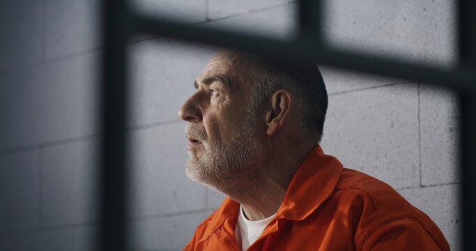 Elderly criminal in orange uniform sits on prison bed and dreams about freedom. Prisoner serves imprisonment term in jail cell. Gloomy inmate in detention center or correctional facility.