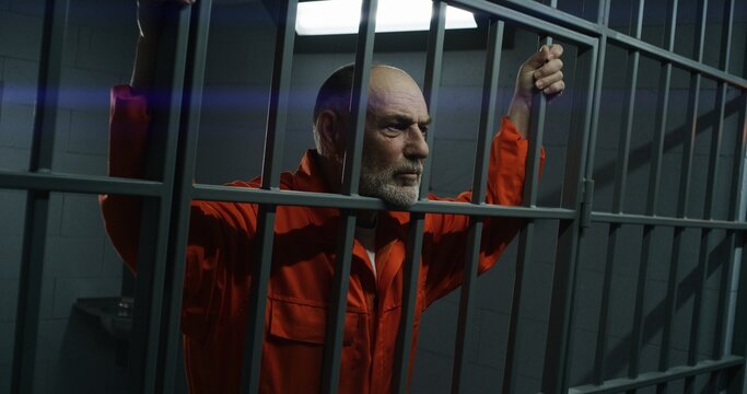 Elderly prisoner in orange uniform stretches fingers, leans on metal bars. Criminal serves imprisonment term for crime in prison cell. Tired inmate stands behind bars in jail or detention center.