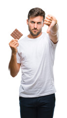 Young handsome man eating chocolate bar over isolated background with angry face, negative sign...
