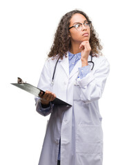 Young hispanic doctor woman holding a clipboard serious face thinking about question, very confused...