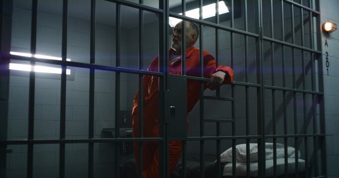Elderly prisoner in orange uniform leans hands on metal bars. Criminal serves term of imprisonment in prison cell. Gloomy inmate stands behind bars in jail, detention center or correctional facility.