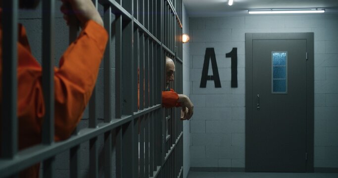 Elderly prisoner in orange uniform stretches fingers, leans on metal bars. Criminal serves imprisonment term for crime in prison cell. Tired inmate stands behind bars in jail or detention center.