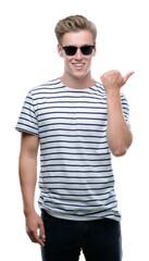 Young handsome blond man wearing sunglasess pointing with hand and finger up with happy face smiling