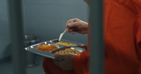 Close up of elderly prisoner in orange uniform sitting in prison cell, eating food. Criminal serves...