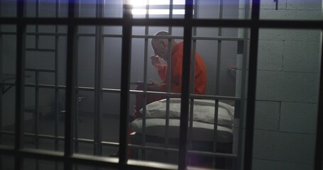 Elderly prisoner in orange uniform sits in prison cell, eats food. Criminal serves imprisonment term for crime. Conditions in jail or correctional facility. Shooting from jail cell through metal bars.