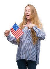 Blonde teenager woman holding flag of United States of America very happy pointing with hand and finger