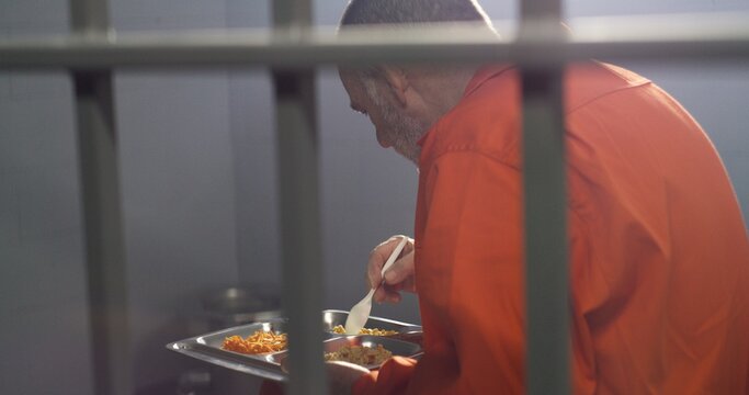 Elderly prisoner in orange uniform sits in prison cell, eats dinner. Criminal, inmate serves imprisonment term for crime. Jail, detention center or correctional facility. Shooting through metal bars.