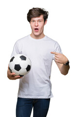 Young man holding soccer football ball over isolated background with surprise face pointing finger to himself