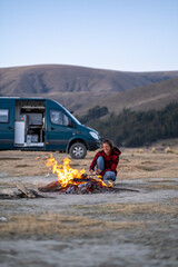 Campsite with a woman in a plaid jacket stoking a fire with a campervan in the background.