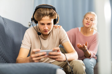 Interested excited teen boy playing online game on cell phone at home while upset mother trying to get his attention. Concept of family phubbing behavior