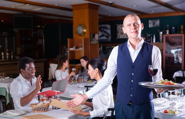 Fototapeta na wymiar Portrait of friendly headwaiter with serving tray warmly welcoming in cozy modern restaurant