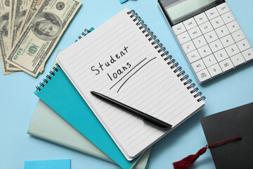 Notebook with text STUDENT LOANS,  calculator, dollar banknotes and graduation cap on blue background