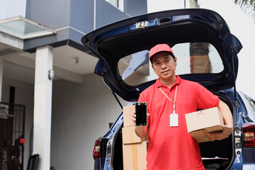 Courier standing near delivery car full of packages outdoors while holding boxes and showing blank...