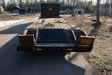 Empty flatbed trailer with raised ramps is parked on the asphalt road in a neighborhood with houses...